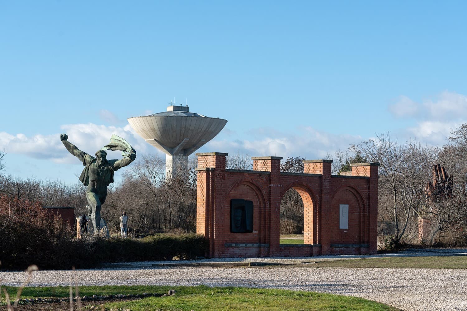 Het bronzen beeld van een aanstormende matroos met vlag naast de rode bakstenen poort van het Memento Park, met op de achtergrond een watertoren onder een blauwe lucht