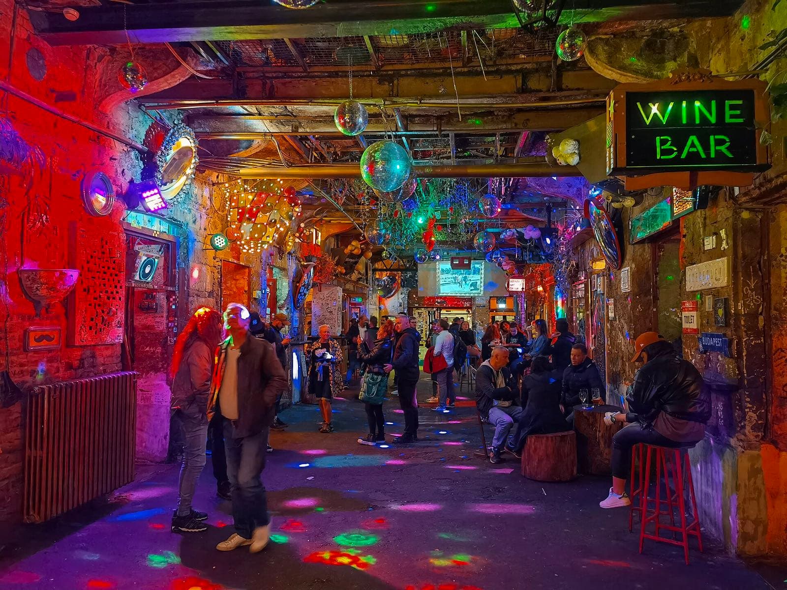 Het kleurrijke interieur van ruïnebar Szimpla Kert met discoballen, neonverlichting, een 'Wine Bar'-bord en bezoekers