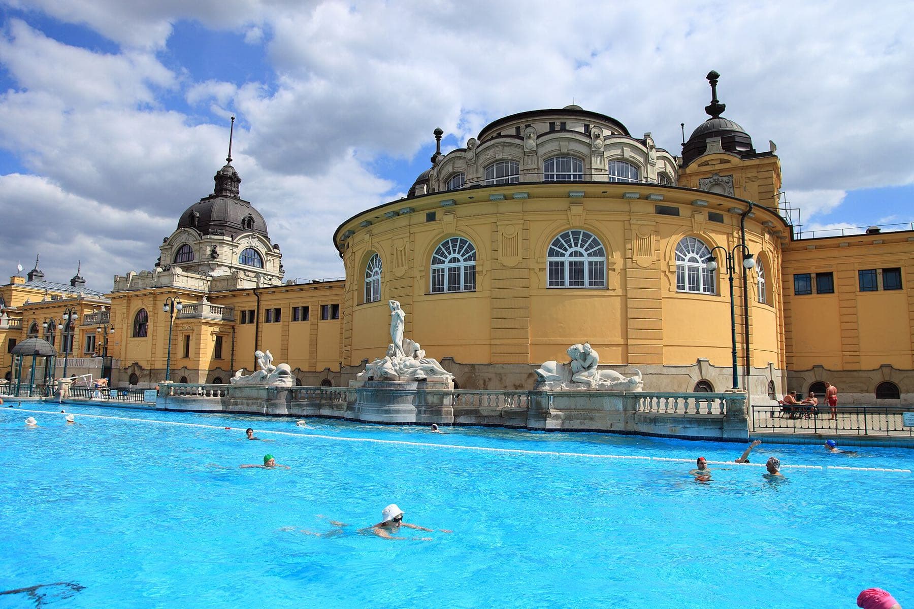 Zwemmers in het helderblauwe buitenbad van het Széchenyi-badhuis, met het gele neobarokke gebouw en koepels onder een wolkenlucht