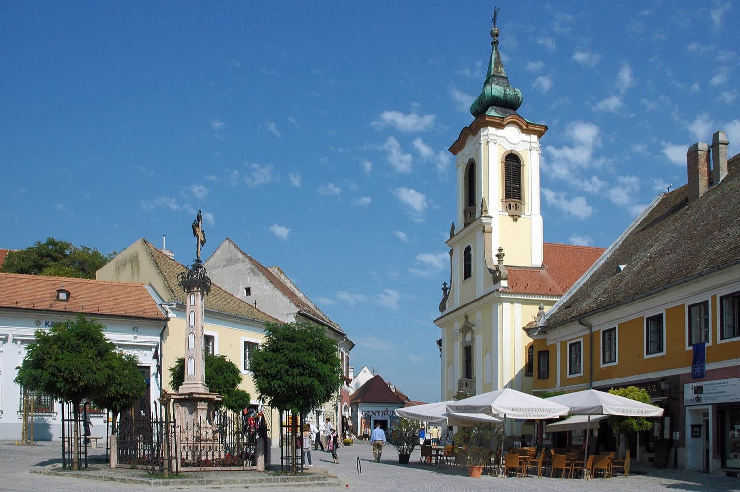 Het Fő tér in het oude centrum van Szentendre met een witte kerktoren, een gedenkkruis, terrasjes en pastelkleurige huizen onder een blauwe lucht