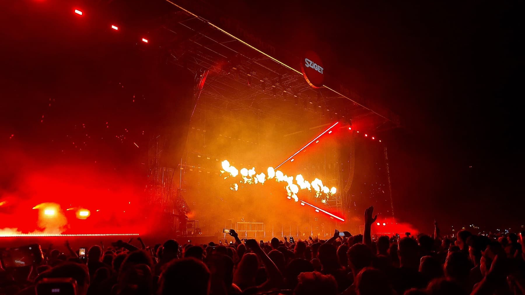 Het hoofdpodium van het Sziget Festival 's nachts, gehuld in rood licht met vuureffecten, gezien over de silhouetten van het publiek